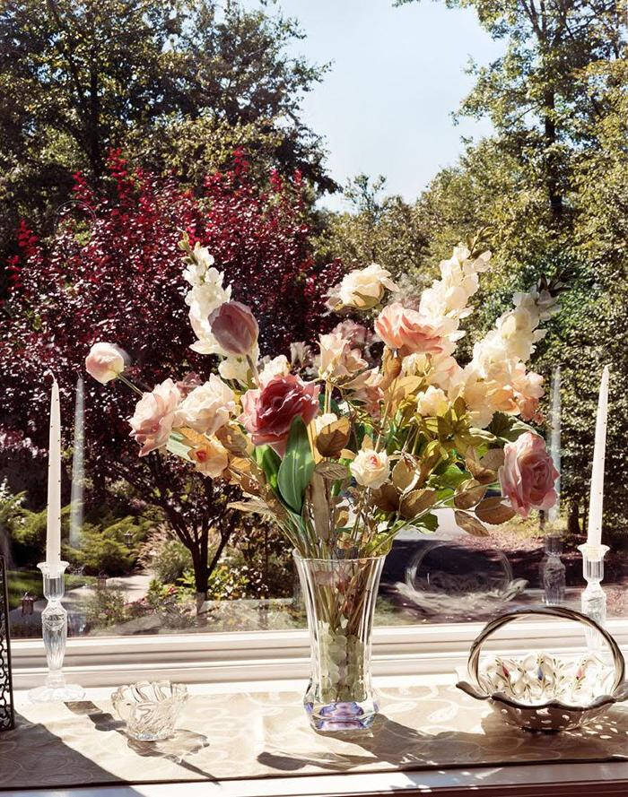 A clear vase of flowers including light pink roses, on a table in front of the window.
